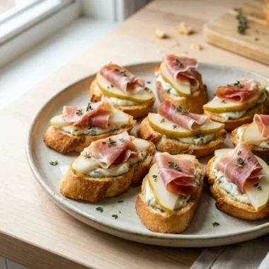 A close-up of a single pear and prosciutto crostini on a white plate.