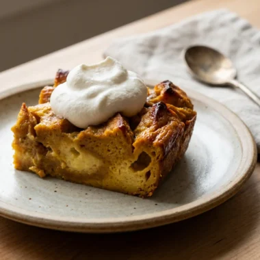 A square slice of pumpkin bread pudding topped with whipped cream on a small plate.